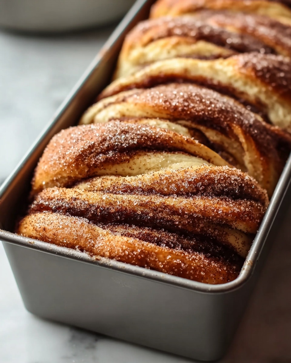 A loaf of pull-apart bread sits in a dark gray baking pan, showing multiple thick layers of golden-brown dough with a soft, fluffy texture. The top layer is sprinkled with a light dusting of sugar and cinnamon, creating a slightly crunchy, textured surface. The bread has deep curved cuts on top, revealing the folded layers underneath, which have a warm, light yellow color with swirls of cinnamon. The pan rests on a white marbled surface that adds a clean, bright contrast to the rich tones of the bread. photo taken with an iphone --ar 4:5 --v 7